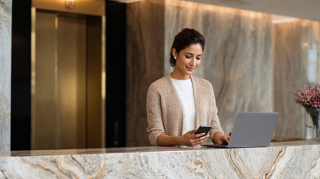 Woman at Reception Desk Working on Laptop and Using Mobile Phone