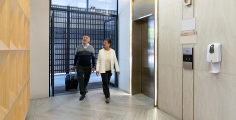 Senior couple enter a hotel lobby through the elevator door. They are dressed casually, carrying luggage, and appear ready to check in and relax after traveling.