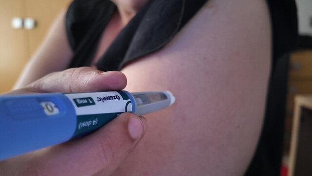 OZEMPIC Woman administering subcutaneous injection. She is using an injector pen on her arm. The pen has a dose selector and view window.