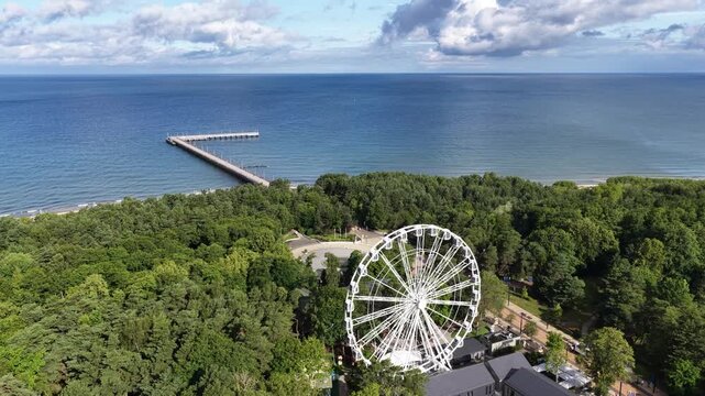 Elevated view of a Ferris wheel rising above lush green trees in Palanga, Lithuania, with a church tower and city buildings visible in the background.
