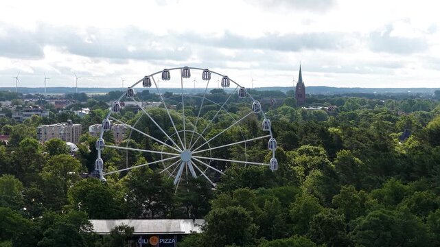 Elevated view of a Ferris wheel rising above lush green trees in Palanga, Lithuania, with a church tower and city buildings visible in the background.