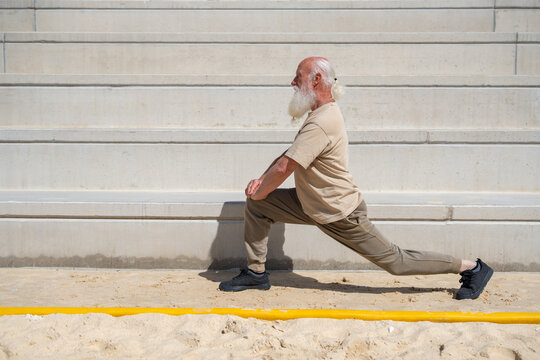 Side view of elderly man with long white beard doing lunge exercise on sandy surface in casual activewear, staying fit and strong outdoors on a sunny day with stone wall background.