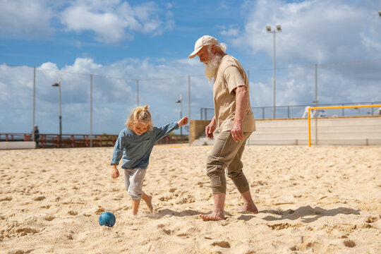 Grandfather and young child playing soccer together on a sandy outdoor field under a bright sky. Multigenerational bonding, active lifestyle, fun family moment, barefoot playtime with a blue ball.