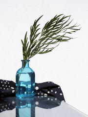 A BoHo-style still life with a sprig of silky oak in a small glass vase and a black and white polka dot napkin on a glass-topped table.