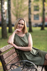 Obraz premium Woman with blonde hair, wearing a green dress, is sitting on a wooden bench in a park, surrounded by trees and soft sunlight, enjoying a peaceful moment outdoors