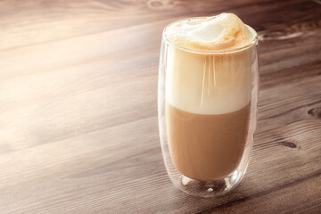 Layered latte in a double-walled glass cup on a wooden surface, showcasing foam, milk, and espresso separation.