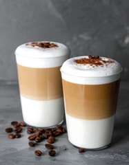 Two layered coffee drinks in tall glasses, topped with foam and cocoa powder, rest on a gray surface scattered with coffee beans