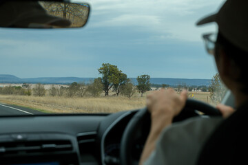 A man wearing glasses driving a car on an open Australian highway surrounded by dry grassland.