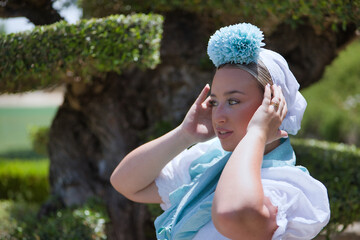 Fototapeta premium Portrait of young woman, beautiful, blonde, with typical Andalusian flamenco costume to go on pilgrimage in white with light blue shawl and flower, placing the headscarf well.