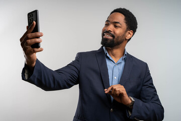 Smiling and cheerful businessman in office taking selfie photo on phone and talking on video call with colleagues and friends using smartphone, african american man waving at camera greeting gesture.