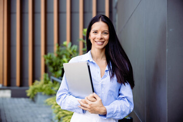 A smiling woman stands outdoors holding a laptop, exuding confidence and approachability in a modern urban environment.