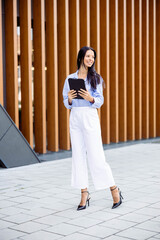 An upbeat woman stands outdoors, holding a tablet as she smiles, representing modern confidence and adaptability amid greenery.