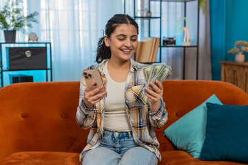 Indian young woman at home sitting on sofa, smartphone in one hand, counting cash dollars with the other. Arabian girl planning personal expenses, budgeting carefully or making online transfers