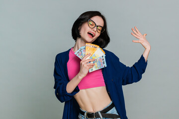 Young woman dances happily and holding money euro cash banknotes in hands, celebrating financial success or good fortune. Girl expresses joyful emotion and enthusiastic celebration on gray background