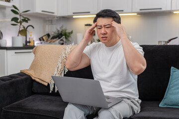 Asian man at home works on laptop feels headache rubs temples closes eyes a moment and breathes slow. Chinese guy on sofa puts laptop aside takes pause does deep breath feels relief and rests quiet