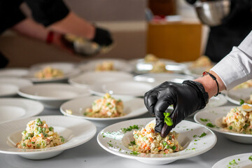 Professional kitchen staff plating vegetable salad on multiple white dishes, wearing gloves for hygiene. Catering preparation with attention to detail and food presentation