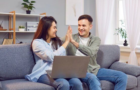 Happy married couple enjoying online, internet leisure activities together at home, using a laptop on sofa. Give each other high five, symbolizing joy and togetherness in the digital age.