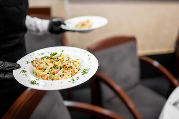 Waiter in black uniform serving two plates of colorful vegetable risotto garnished with herbs in a restaurant setting. Professional food presentation and service