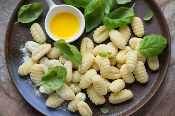 Raw potato gnocchi with flour, green basil leaves and olive oil on a brown plate, horizontal shot, middle close-up