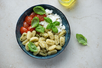 Bowl of potato gnocchi with ricotta, roasted tomatoes and fresh basil, horizontal shot on a light-beige stone background, top view