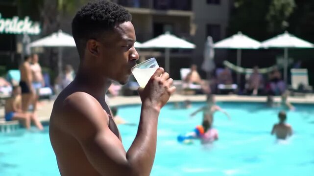 shirtless young african american man with drink relaxes by crowded swimming pool on sunny day. summer vacation and resort lifestyle concept. travel and leisure, tourism and hospitality
