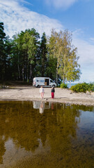 Two individuals stand at the edge of Lake Saimaa, absorbing the serenity of the landscape. A camper van rests nearby, surrounded by vibrant trees under a blue sky, creating a picturesque scene.