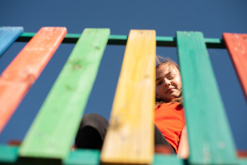 A happy child looking down through colorful wooden playground slats against clear blue sky, playful perspective. Enjoying a fun and active outdoor moment
