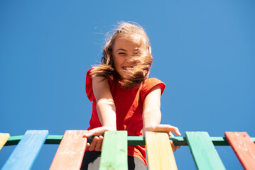 A bright, smiling child girl in a red shirt plays on a colorful wooden playground against a clear blue sky, with her windy hair covering her face, enjoying a fun and active outdoor moment