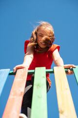 A bright, smiling child girl in a red shirt plays on a colorful wooden playground against a clear blue sky, with her windy hair covering her face, enjoying a fun and active outdoor moment