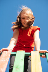 A bright, smiling child girl in a red shirt plays on a colorful wooden playground against a clear blue sky, with her windy hair covering her face, enjoying a fun and active outdoor moment