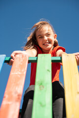 A smiling child in red shirt playing on colorful wooden playground against clear blue sky, enjoying a fun and active outdoor moment.