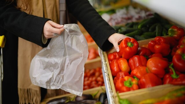 Woman's hands picking fresh red bell pepper from a produce display into a plastic bag at a supermarket