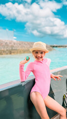 Portrait of an 8-year-old girl in a swimsuit and hat smiling on a boat near a tropical beach. Bright daylight, cheerful mood, summer vacation atmosphere. Kids summer activity