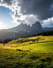 Sunlit meadow and rolling hills below dramatic, imposing mountains under a partly cloudy sky