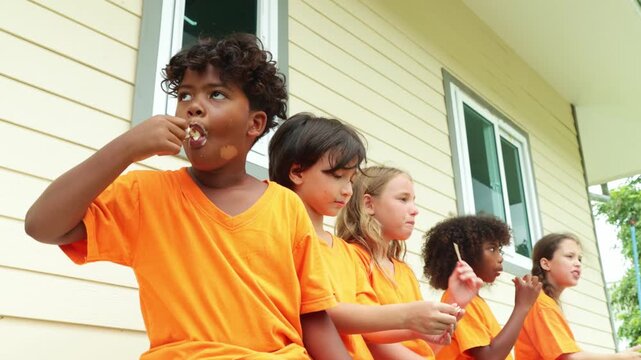 Group young male and female international school students sit in a row eating popsicles to cool down and enjoy their favorite delicious cool ice cream after playing outdoor sports during the summer.