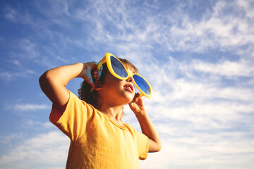 A happy little boy wearing large, funny sunglasses enjoys the sun. The boy stands against a blue sky on a sunny day