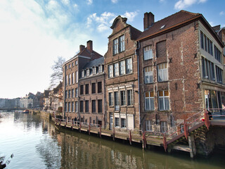 Naklejka premium Riverside historic houses in Ghent, Belgium - A row of traditional brick buildings with reflections in the calm water of the canal on a sunny day in Ghent.