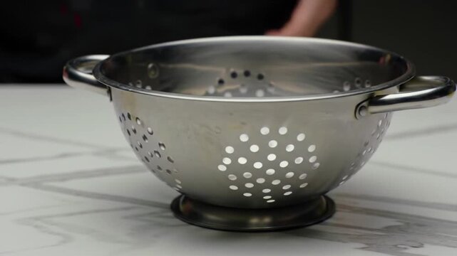 Stainless steel colander on a marble countertop with hands preparing to drain pasta in a kitchen setting