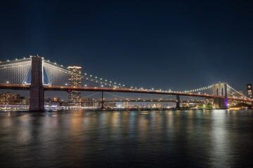 Obraz premium A night view of a cityscape New York with illuminated buildings, Brooklyn bridge in the foreground, and a cloudy sky. Real photo, , 4k