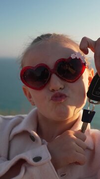 Child car keys heart sunglasses, cute girl makes kissy face holding car keys. Vertical video.