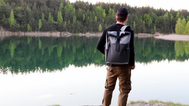 A man stands by a calm lake, organizing his gear before heading out to fish. The area is peaceful, with trees lining the shore and the water reflecting the soft morning light