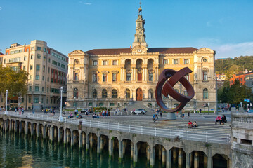 Bilbao City Hall and riverside sculpture. - The historic Bilbao City Hall and its prominent sculpture by the Nervión River, with people enjoying a sunny day on the waterfront.