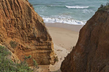 coastal view with golden cliffs, sandy beach, and turquoise ocean waves. The cliffs feature layered rock and sparse vegetation, with buildings visible on top in Portimao, Algarve, Portugal © hectorchristiaen