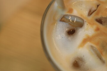 Iced coffee sits in a clear glass, featuring cream and foam layered above visible ice cubes. The setting is a rustic wooden table, creating a warm atmosphere.