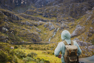Latino man wearing hoodie and backpack exploring Santurban moor in Colombia, standing near a lagoon surrounded by rocky mountains, symbolizing hiking, eco travel, adventure, freedom and nature.
