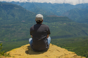 Latino man sitting at the edge of a viewpoint in Barichara Colombia, looking at the mountains, representing freedom, mindfulness, travel, nature connection, adventure, peace, exploration and lifestyle