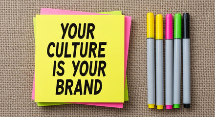 Yellow sticky note with text "Your Culture Is Your Brand", stacked on pink and green notes, beside colorful markers on burlap background, showcasing a branding concept