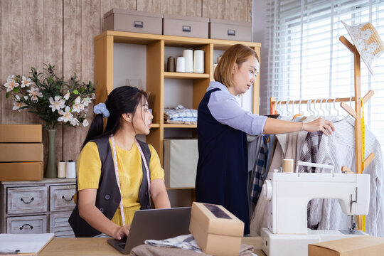 Two female partners running clothing shop together, one using phone and laptop to receive order while other checking garments, teamwork showing cooperation in small business startup fashion store