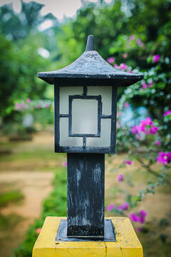 Foto close-up sebuah lampu taman dengan desain kotak klasik berwarna hitam, dipasang di atas tiang kayu berwarna kuning. Latar belakang berupa pepohonan hijau dan suasana taman yang tampak blur (bokeh
