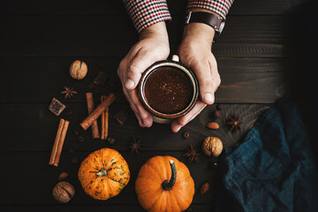 Top view of male hands holding mug of hot chocolate, pumpkins and spices on rustic wooden background, cozy autumn scene